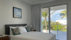 A bedroom with a bed, a nightstand, a framed picture on the wall, and a view of palm trees and a building through the window.
