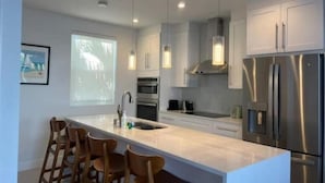 A modern kitchen with a large island, stainless steel appliances, pendant lighting, and a projection screen on the wall.