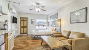 A compact room with a bed, a sofa, a wooden table, a ceiling fan, a TV, and a window with a view of buildings.