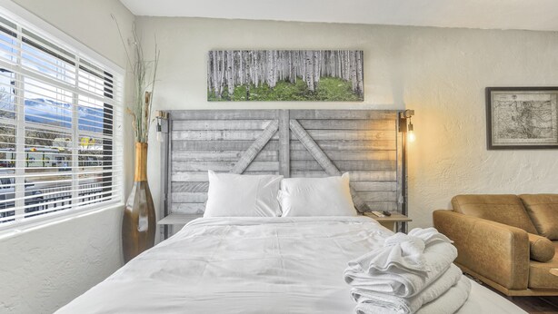A bedroom with a bed, a wooden headboard, a window with blinds, a vase with plants, a map on the wall, and a leather armchair.
