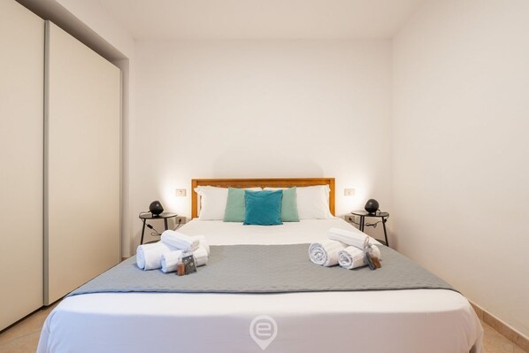 A neatly made bed with white linens, a grey blanket, and two teal pillows. There are towels and toiletries on the bed.