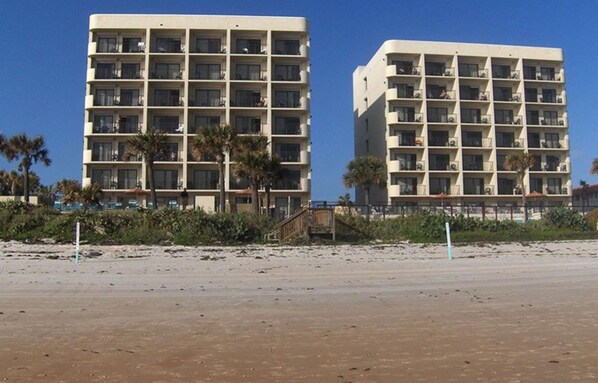Two multi-story buildings with balconies, located near a sandy beach with palm trees.