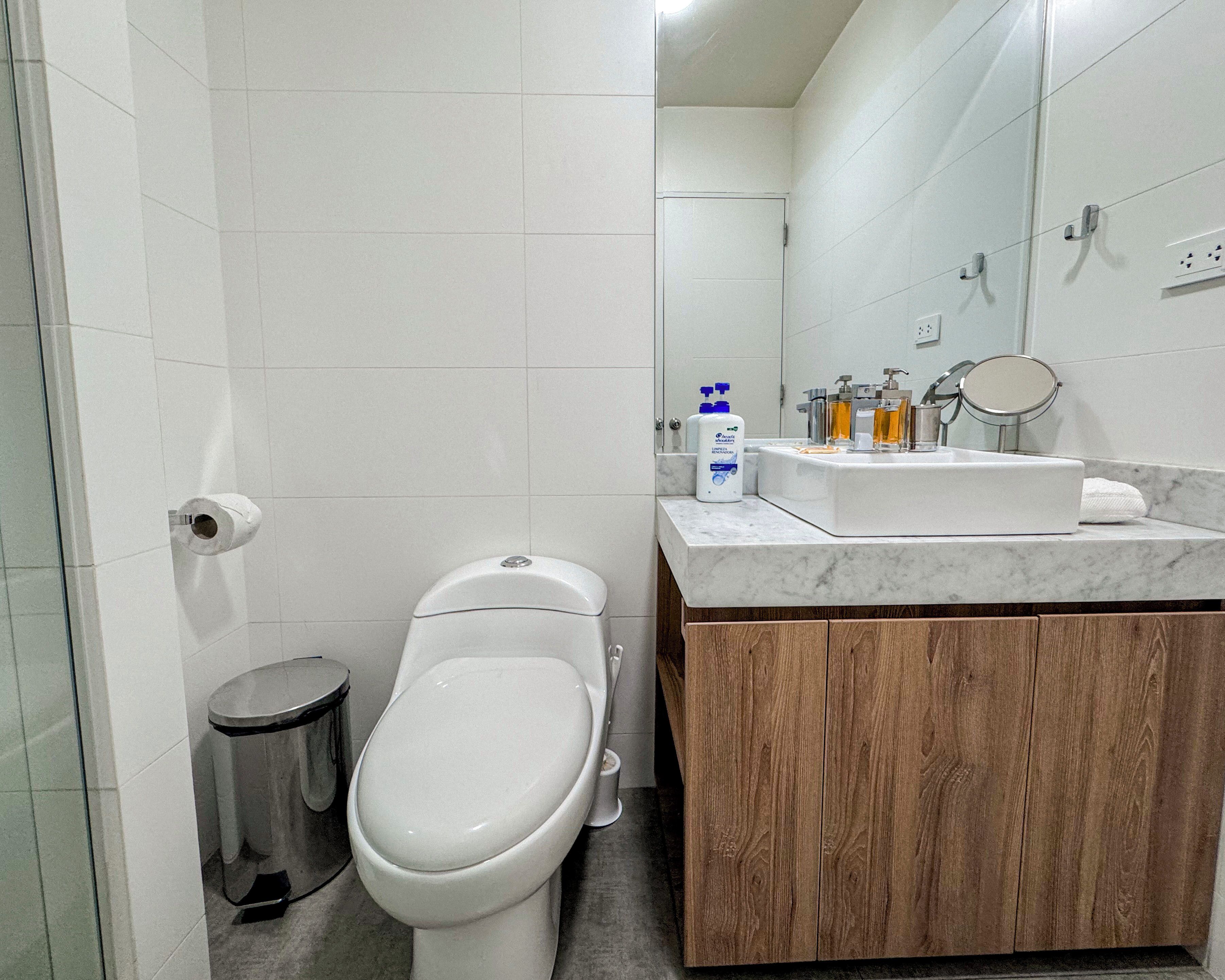 Un baño moderno con inodoro blanco, un bote de basura, lavamanos de mármol y tocador de madera.