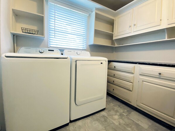 A laundry room with two white washers and dryers, a window with blinds, and white cabinetry with open shelves.