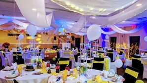 Een feestzaal met ronde tafels, ingericht voor een formeel evenement, met witte en gele tafelkleden, ballonnen en lichtslingers.
