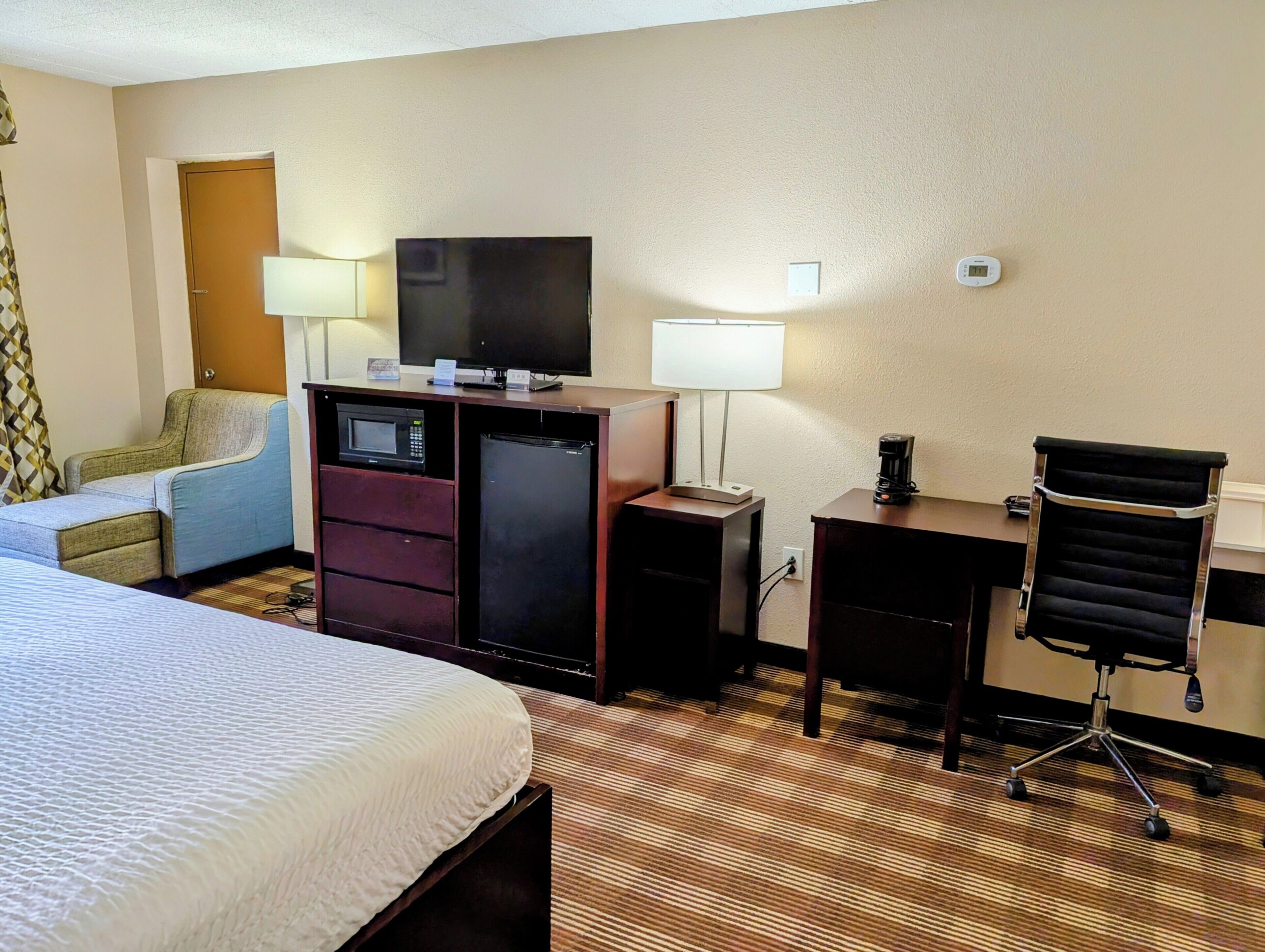 A hotel room with a bed, a desk with a computer, a microwave, a refrigerator, a chair, a TV, and a coffee maker.