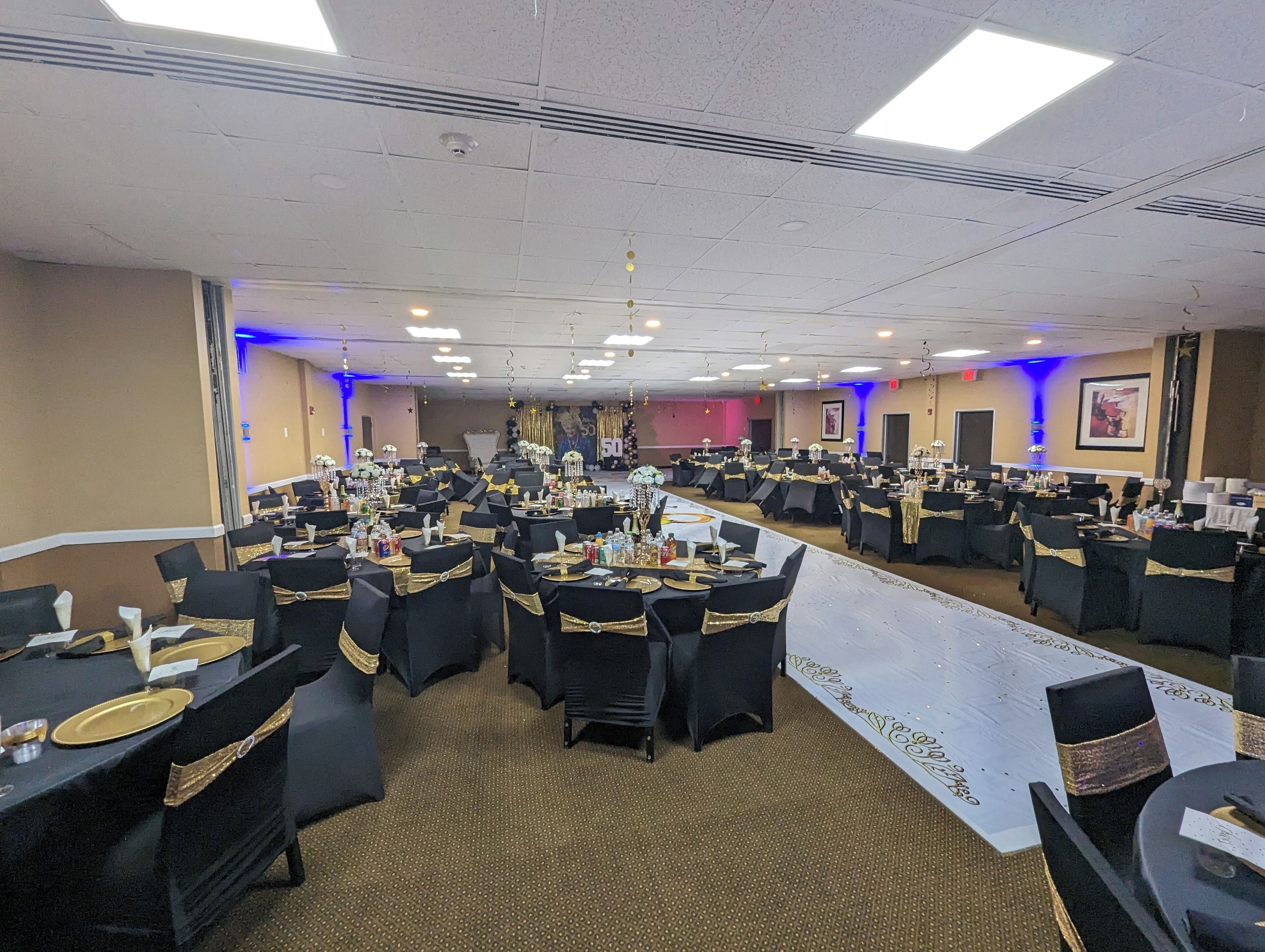 A large banquet hall with round tables, black chairs with gold sashes, and a stage at the front.