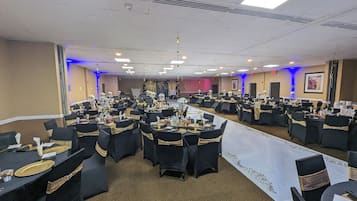 A large banquet hall with round tables, black chairs with gold sashes, and a stage at the front.