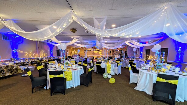 Een feestzaal met gedekte tafels voor een formeel evenement, bedekt met witte stof en gele linten, verlicht door lichtslingers.