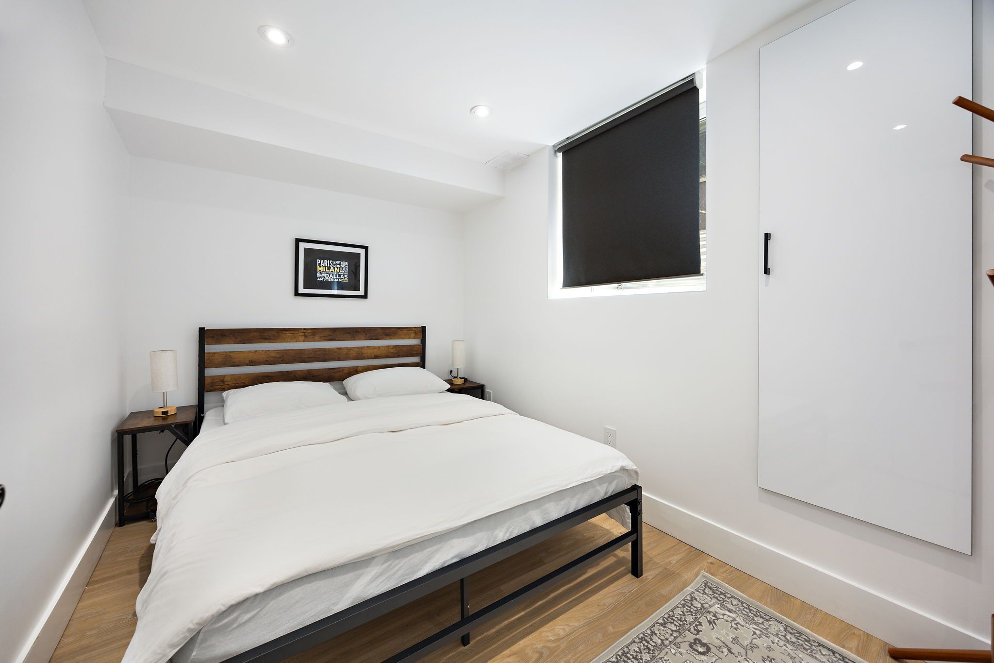 Une chambre à coucher moderne avec un cadre de lit en bois, du linge de lit blanc, une table de chevet avec une lampe, un tableau encadré au mur et une grande armoire.