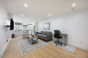 Un salon moderne avec un canapé gris, un fauteuil de bureau noir, une table à manger avec des chaises, un téléviseur fixé au mur et un coin cuisine à l’arrière-plan.