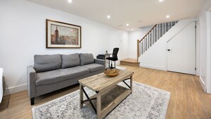 A living room with a grey sofa, a wooden coffee table, a framed cityscape painting, and a staircase with a wooden railing. - Cozy & Charming Apt For 4 | Quiet & Privacy (Pickering)
