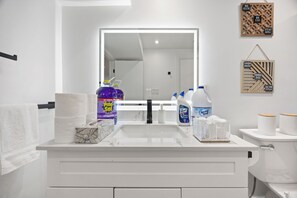 Une salle de bain avec un lavabo blanc, un miroir et des toilettes. On trouve des produits de nettoyage sur le lavabo, notamment un grand flacon de Pine-Sol, un flacon d’Ajax et un flacon de Spring Home. Il y a également deux rouleaux de papier toilette sur les toilettes.