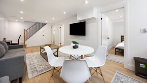 A modern living room with a gray sofa, white chairs, a round table with a bonsai, a TV on the wall, and a staircase. - Cozy & Charming Apt For 4 | Quiet & Privacy (Pickering)