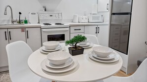 A modern kitchen with white cabinetry, a stove, microwave, and refrigerator. There is a dining table with white plates and a bonsai tree centerpiece. - Cozy & Charming Apt For 4 | Quiet & Privacy (Pickering)