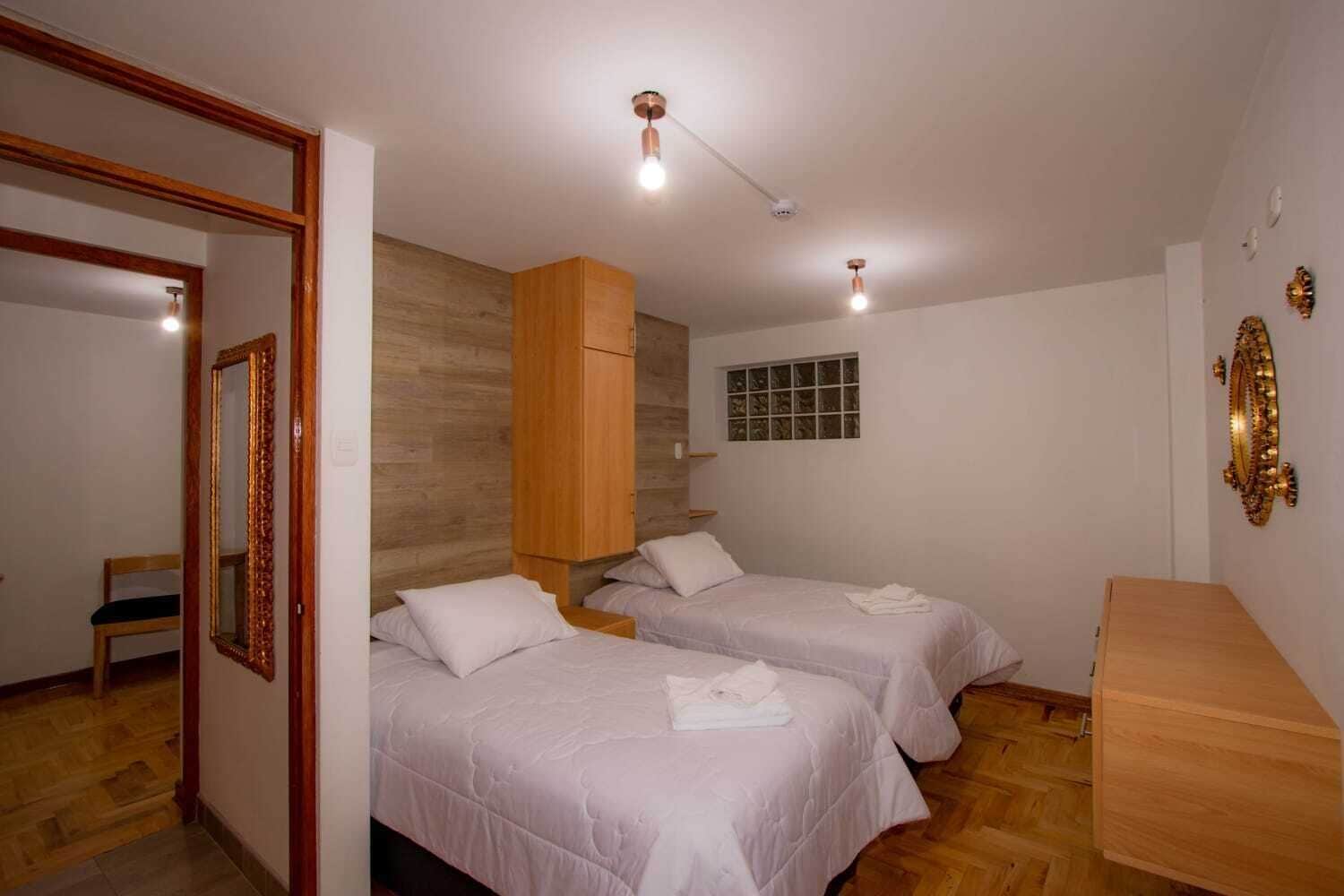 A hotel room with two beds, a wooden wardrobe, a mirror, and a small window.