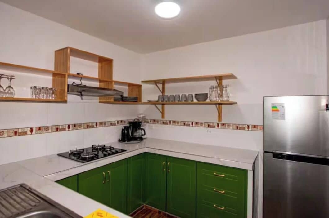 A kitchen with green cabinets, a stainless steel refrigerator, and wooden shelves above the counter.
