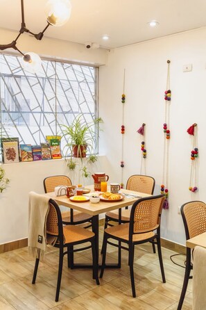 A dining area with a table set for breakfast, wicker chairs, a potted plant, and a wall with colorful decorations.