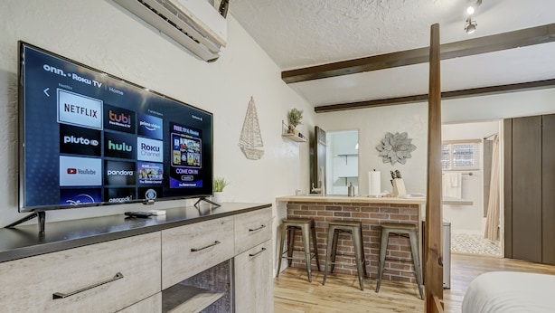 A modern hotel room with a large flat-screen TV displaying streaming services, a wooden floor, a bed, a small kitchen area with stools, and a mirror on the wall. - Quest Rest - Unit 6 (Colorado Springs)