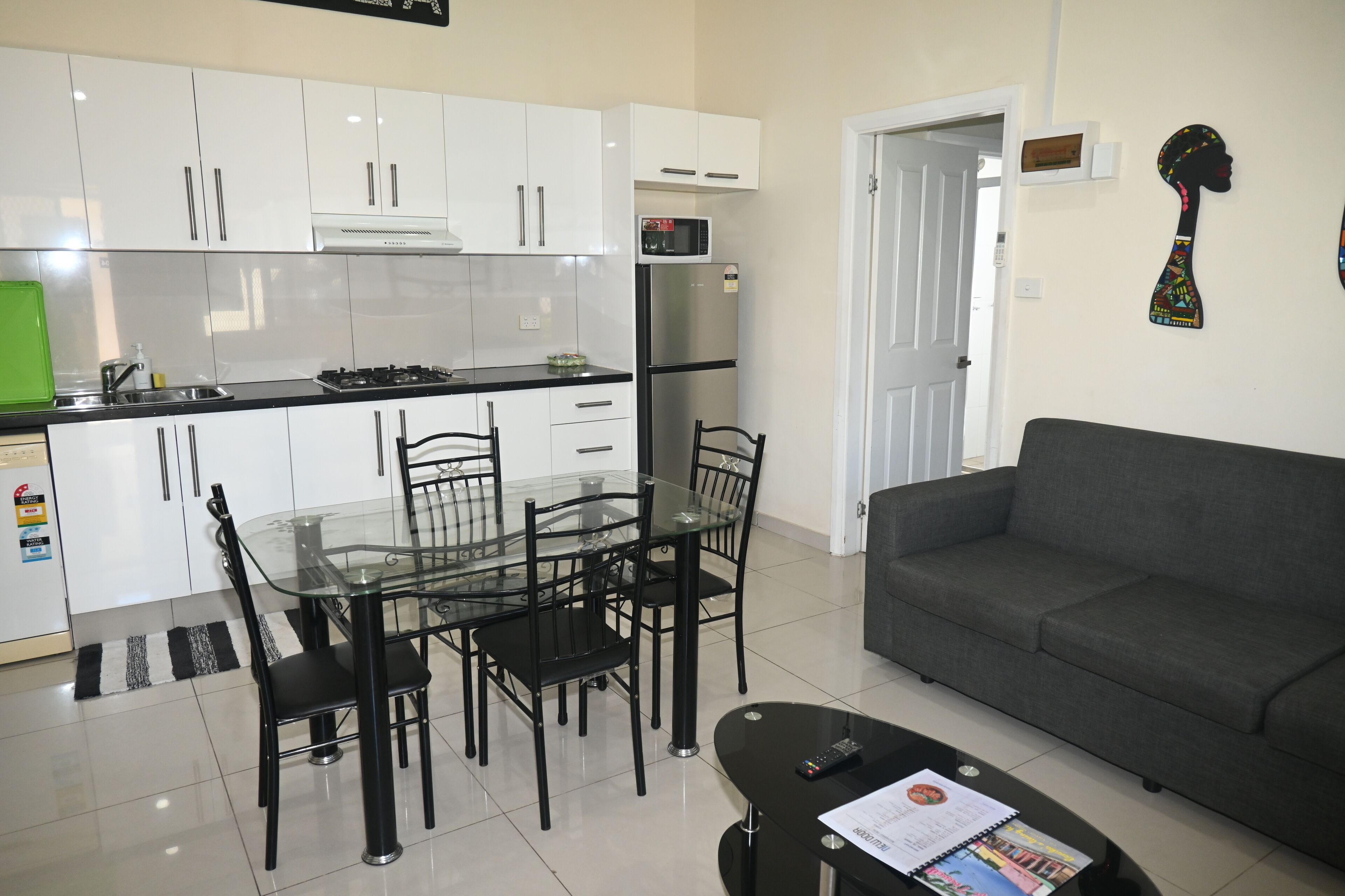 A modern kitchen with white cabinets, a black refrigerator, and a microwave. There is a dining table with chairs and a grey sofa in the living area.