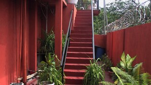 A red building with a staircase leading to a balcony, surrounded by potted plants and a palm tree.