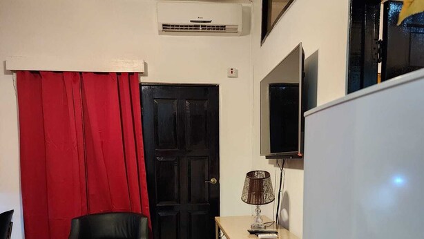 A compact living space with a black door, a red curtain, a black leather chair, a small table with a lamp, and a white refrigerator.