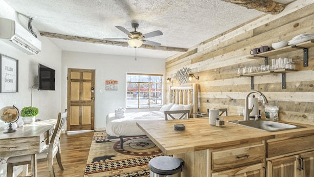 A rustic kitchen with wooden cabinets, a sink, and a ceiling fan. There is a dining area with a table and chairs, and a bed in the background.