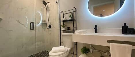 A modern bathroom with a glass shower enclosure, a toilet, a vanity with a mirror, and a small shelf with toiletries.