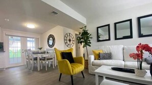 A modern living room with a yellow armchair, white sofa, and a coffee table. There is a dining area with white chairs and a table in the background.