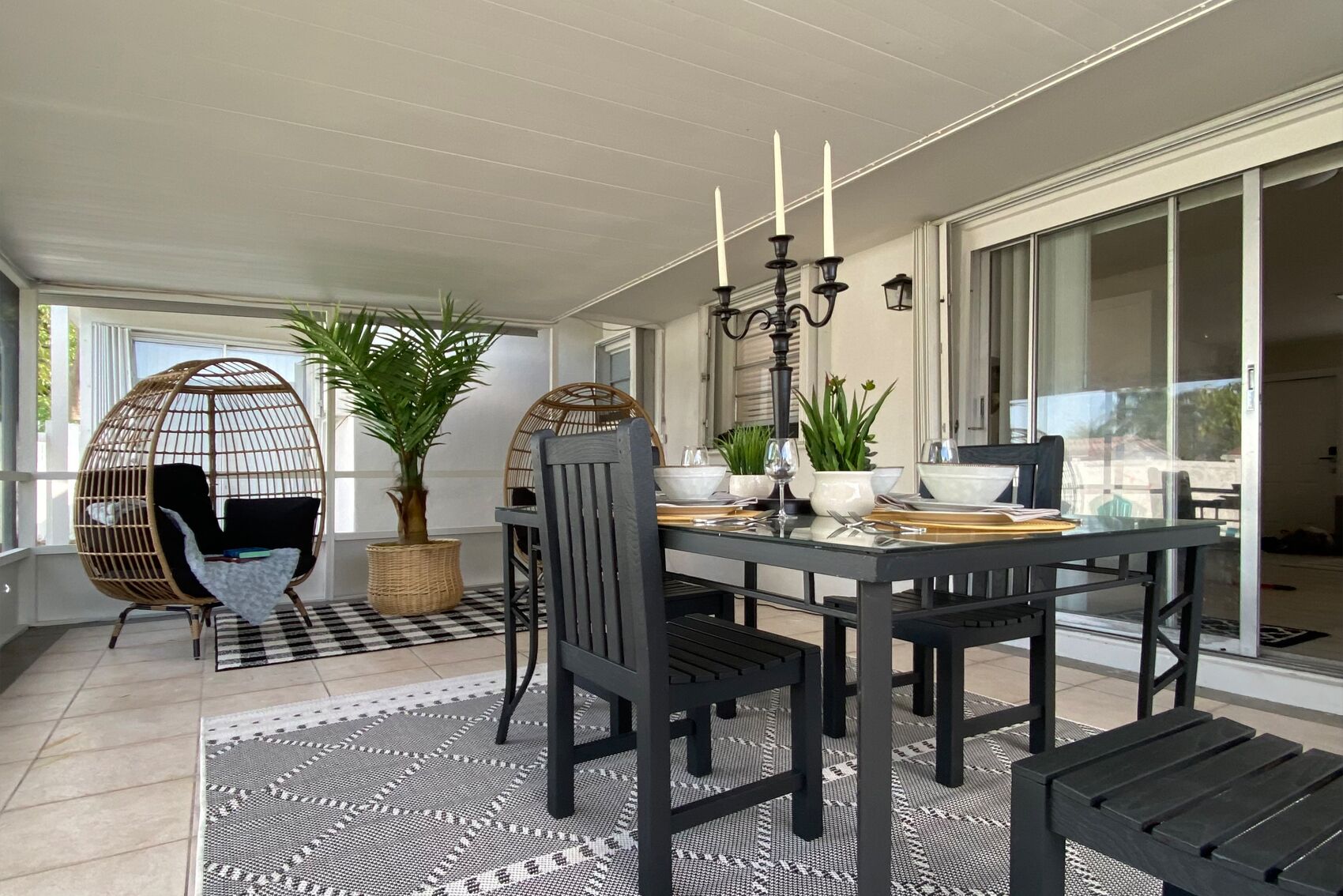 A dining area with a black table set for four, surrounded by black chairs. There is a wicker chair with a cushion, a potted plant, and a candelabra on the table.