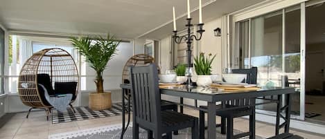 A dining area with a black table set for four, surrounded by black chairs. There is a wicker chair with a cushion, a potted plant, and a candelabra on the table.