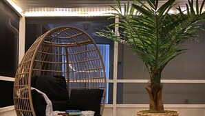 Una habitación con un ventanal grande, una silla de mimbre con cojín negro, un tapete blanco y mullido, y una planta en maceta.