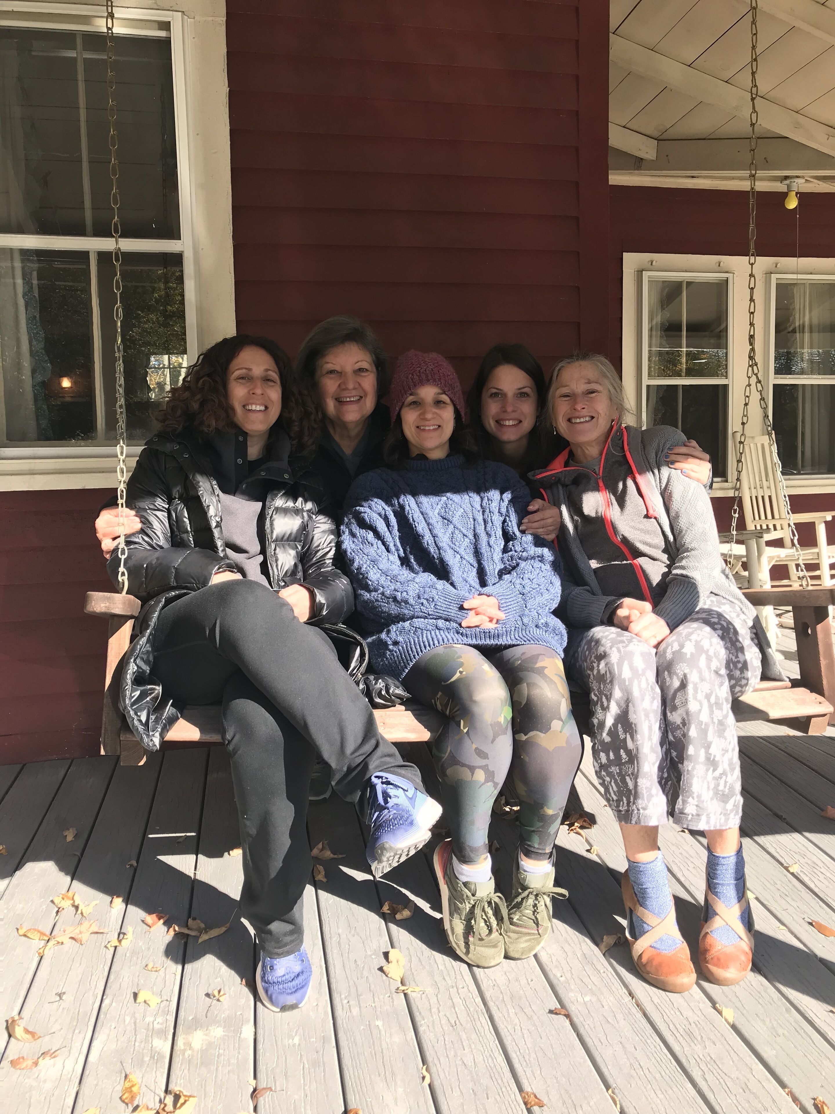 A group of people sitting on a porch with a swing, wooden floor, and a window with a view of trees.