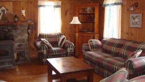A cozy living room with plaid furniture, a stone fireplace, and a wooden bookshelf. - Farmhouse on Otis Pond (Otis)