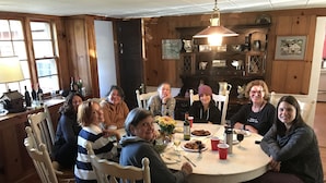 A group of people seated around a round dining table in a room with wooden paneling and a chandelier. - Farmhouse on Otis Pond (Otis)