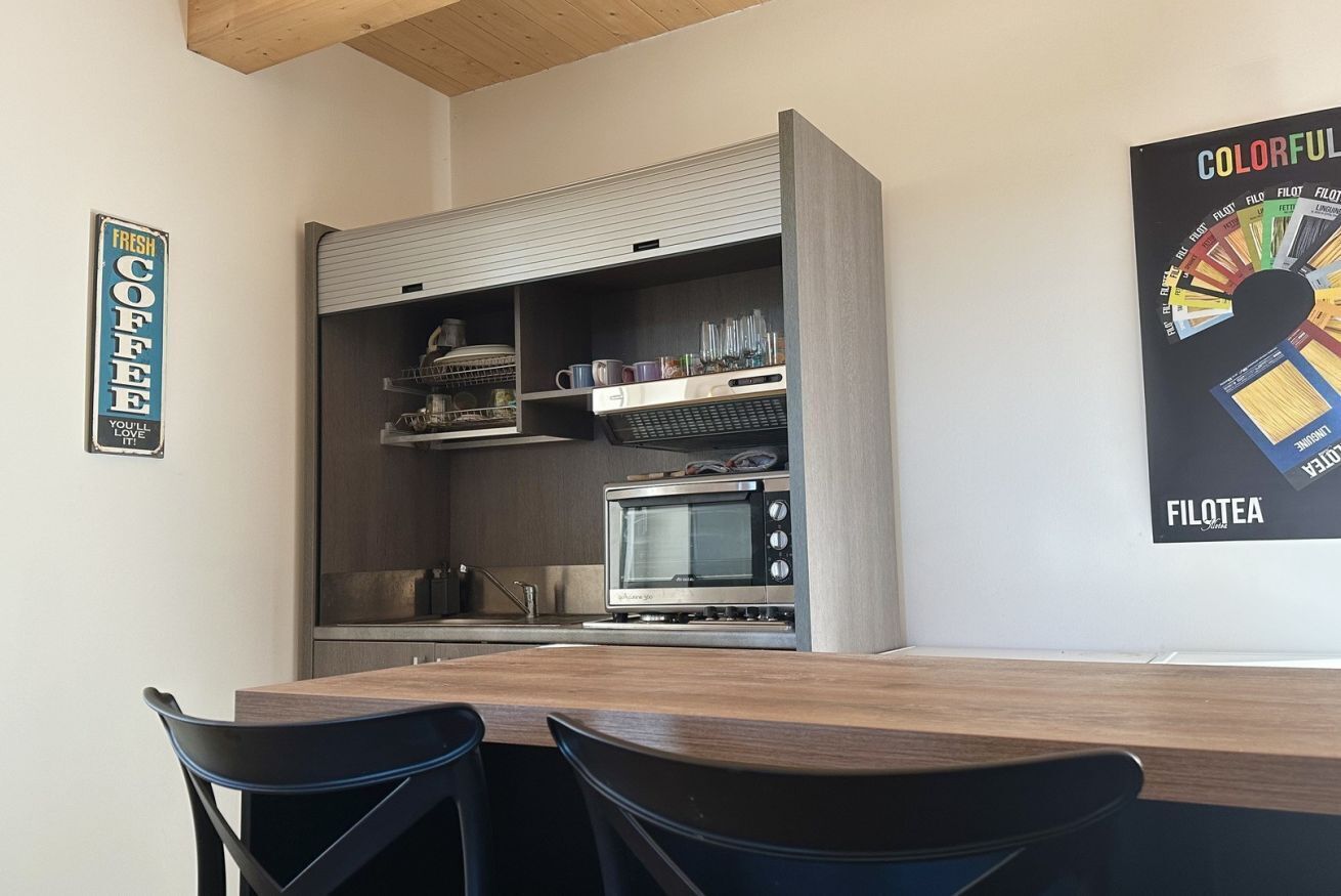 A compact kitchen with a microwave, toaster, and coffee machine, a wooden dining table, and chairs.