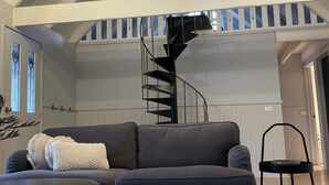 Un loft espaiós amb un sostre alt, una escala de caragol, un sofà gris, una taula de centre rodona i una catifa amb un disseny original.