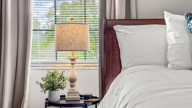 A neatly made bed with white linens, a wooden bedside table with a lamp, a potted plant, and a tray with a cup and a book. - Family Friendly with Kid activities (Tifton)