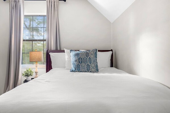 Une chambre avec un grand lit, une fenêtre avec des stores, une lampe et une petite plante.