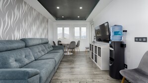 Una sala de estar moderna con un sofá gris, una mesa de comedor con sillas, un televisor de pantalla plana, un dispensador de agua y vistas al exterior a través de amplios ventanales.