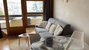 A modern living room with a glass table, white chairs, a grey sofa, and a view through large windows.