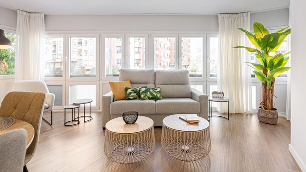 Ein modernes Wohnzimmer mit einer Couch, zwei runden Tischen, einer großen Pflanze und Blick auf Gebäude durch große Fenster.