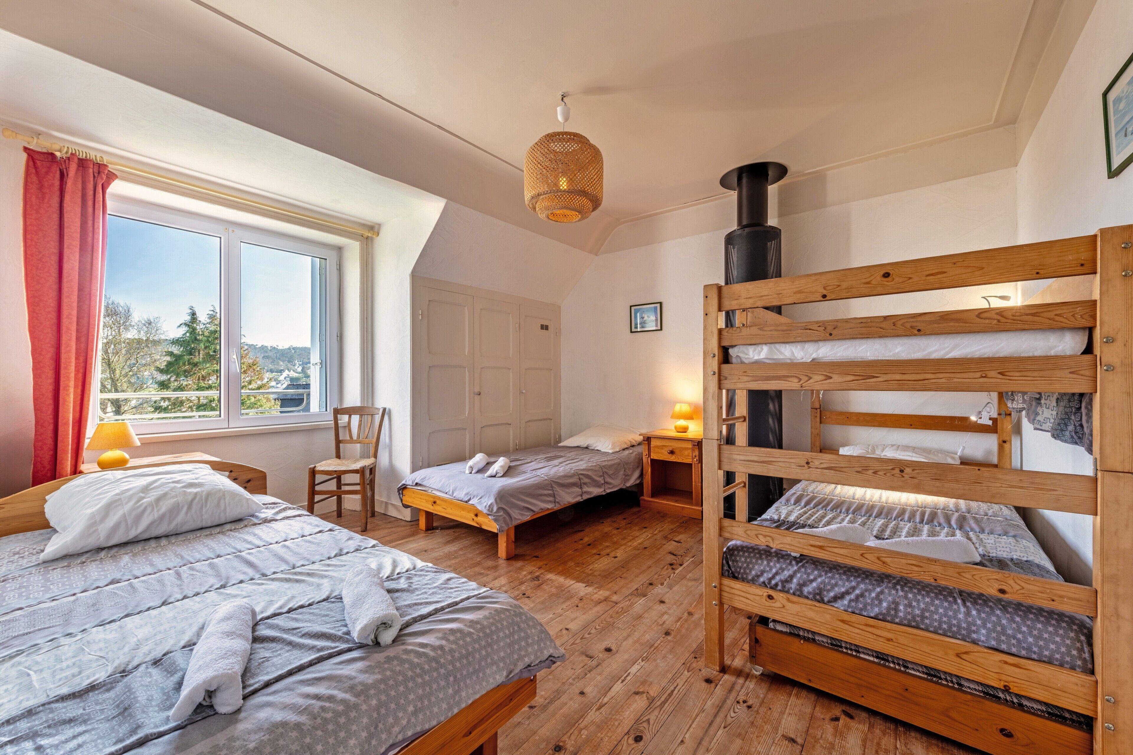 Een kamer met twee bedden, een stapelbed van hout, een raam met gordijnen, een stoel, een tafel met een lamp en een wandradiator.