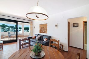 Uma sala de jantar com mesa de madeira, cadeiras, luminária pendente e vista para uma área de estar ao ar livre através de uma porta de vidro.