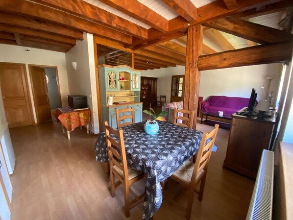 Un comedor con una mesa, sillas y un jarrón azul. Se aprecia un techo de madera y un sofá morado al fondo.