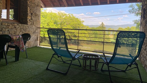 Una terraza con pared de piedra, techo de madera, dos sillas verdes, una mesita redonda y vistas a árboles y montañas verdes.