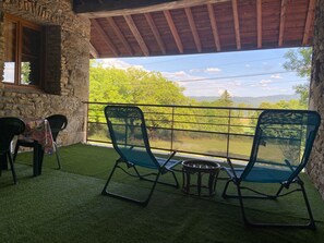 A terrace with a stone wall, a wooden ceiling, two green chairs, a small round table, and a view of green trees and mountains.