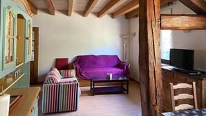 Una habitación con techo de madera, un sofá morado, un reposapiés a rayas, una mesita y un televisor.