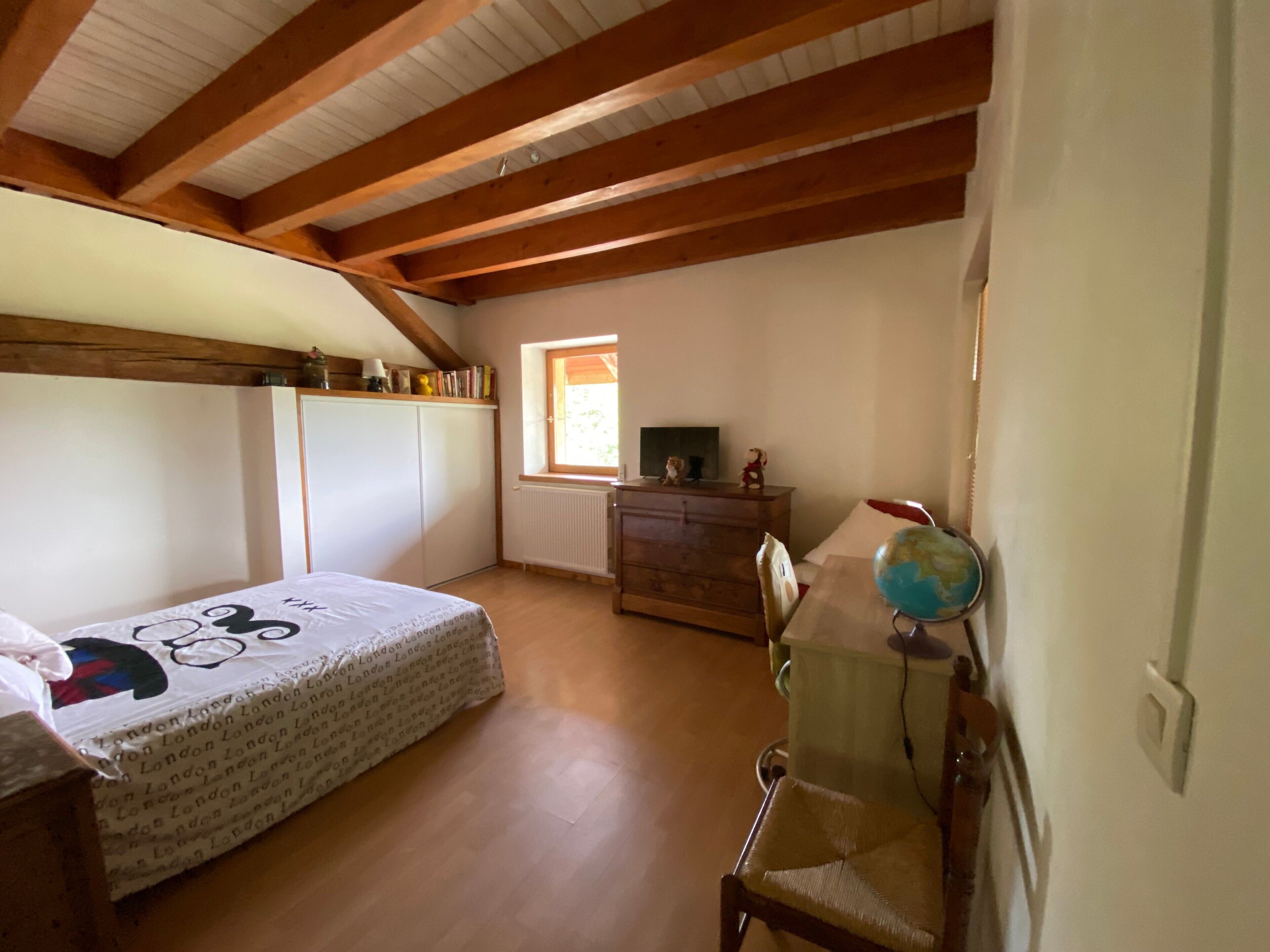 A bedroom with a wooden ceiling, a bed with a patterned bedspread, a dresser, a desk with a globe, and a window with a view.