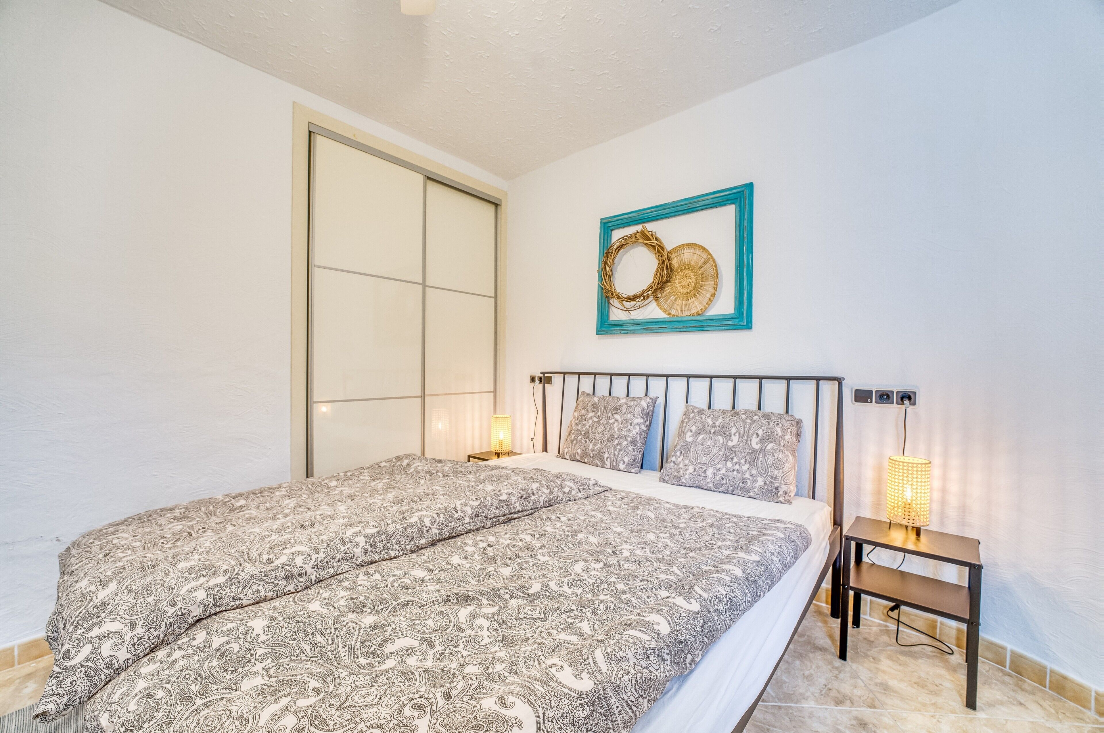 A bedroom with a bed, a nightstand, a wall-mounted light, and a decorative wall piece.